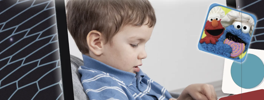 The image shows a young child, dressed in a blue striped shirt, focused on using a tablet or similar device. In the upper right corner of the image, there is a small icon featuring Elmo and Cookie Monster, characters from Sesame Street, suggesting that the child might be interacting with a Sesame Street app, possibly "Sesame Street Alphabet Kitchen." The background is plain, keeping the focus on the child and the device